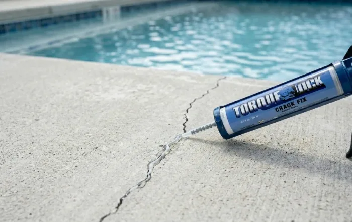 using a caulk gun to inject clear low-viscosity epoxy into a hairline crack in a light gray concrete pool deck. Natural daylight, shallow depth of field, soft shadows. A cartridge labeled 'Crack Fix Epoxy' is visible in the foreground. Clean, modern, trustworthy look. Shot from a slight overhead angle. Blue pool water softly out of focus in the background to suggest pool repair context.